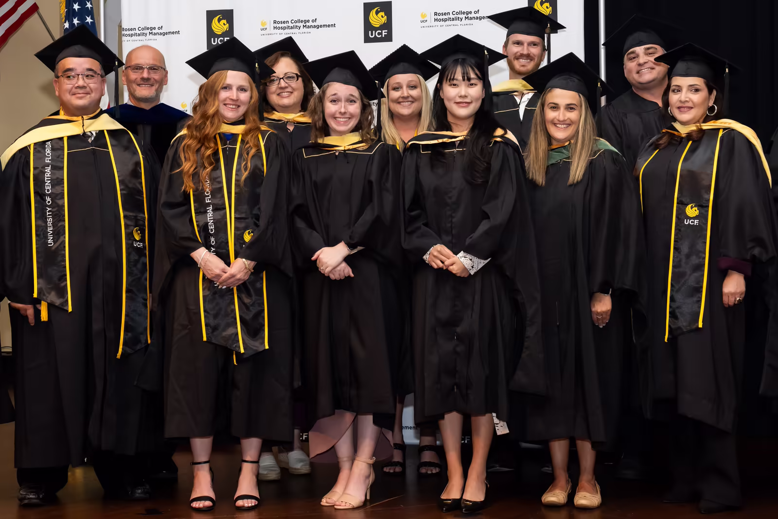 Graduate students in black gowns with gold hoods posing on stage at a hooding ceremony