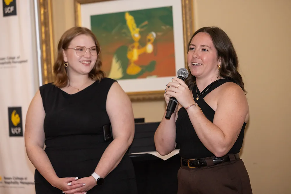 Two women in black speaking into a microphone in front of a UCF Rosen College banner