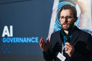 A man wearing glasses speaks into a microphone with a screen behind him displaying "AI GOVERNANCE."