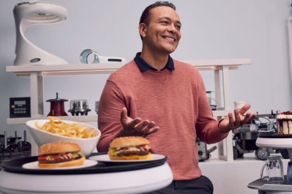 Interview with Juan Higueros, Bear Robotics COO, smiling and gesturing in a kitchen setting with hamburgers and fries on a tray of a service robot.